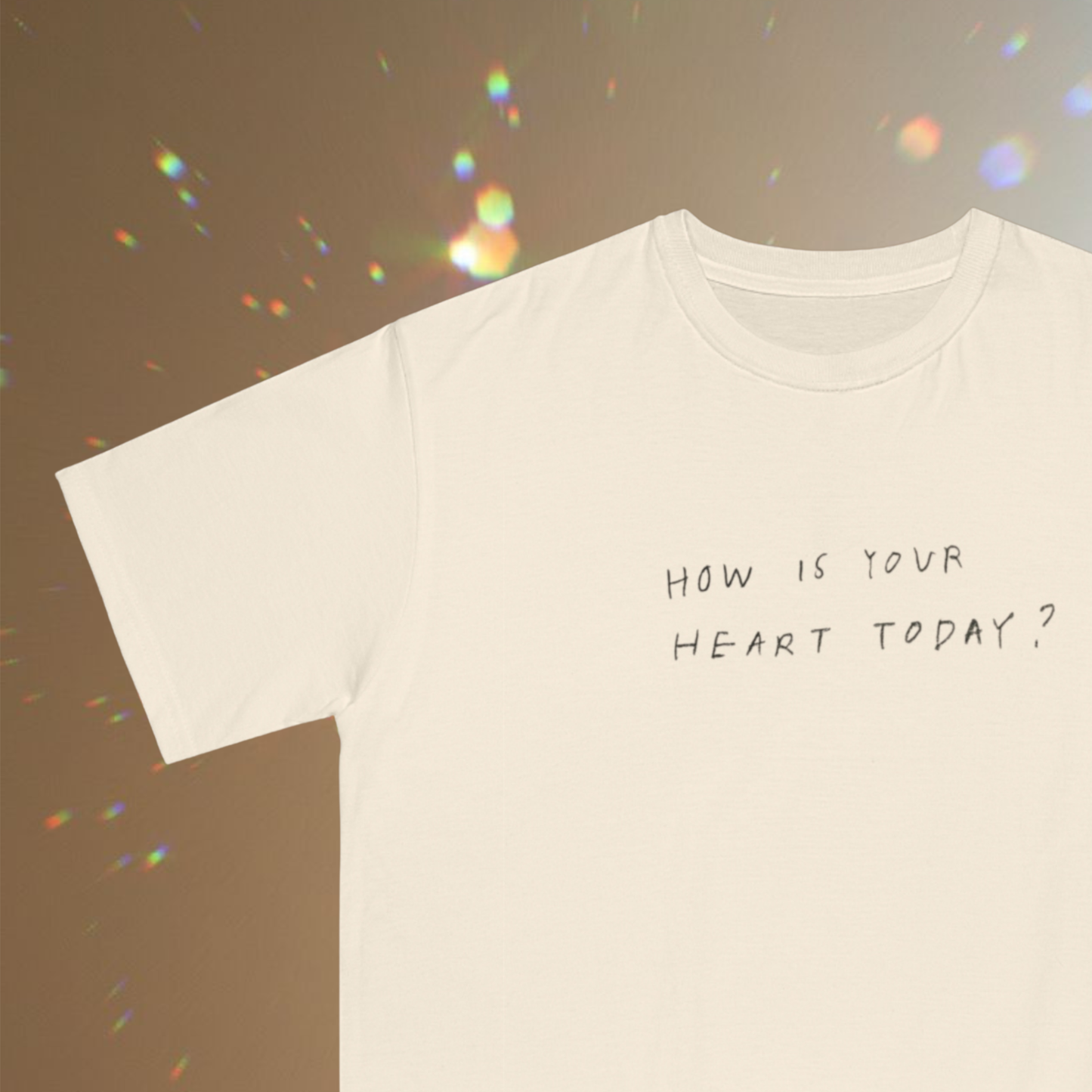 trendy t-shirt with quote hand written ' how is your heart today?' in black and small presentation of the t-shirt on a faded brown background with points of multi color light  