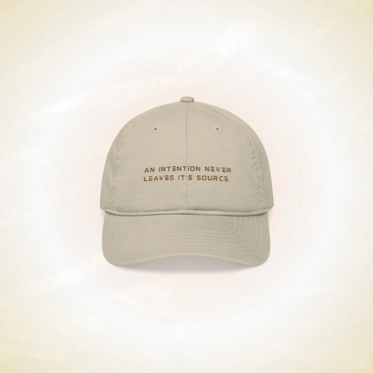 beige baseball cap with the statement ' an intention never leaves it's source' on a ligh background representing protection shield designed spiritually this hat is a crown chakra protection protecting from mental interferences, distractions, fatigue, negatives energies, innovative spiritual fashion concept for spiritual people, a solution to spiritual people problems 